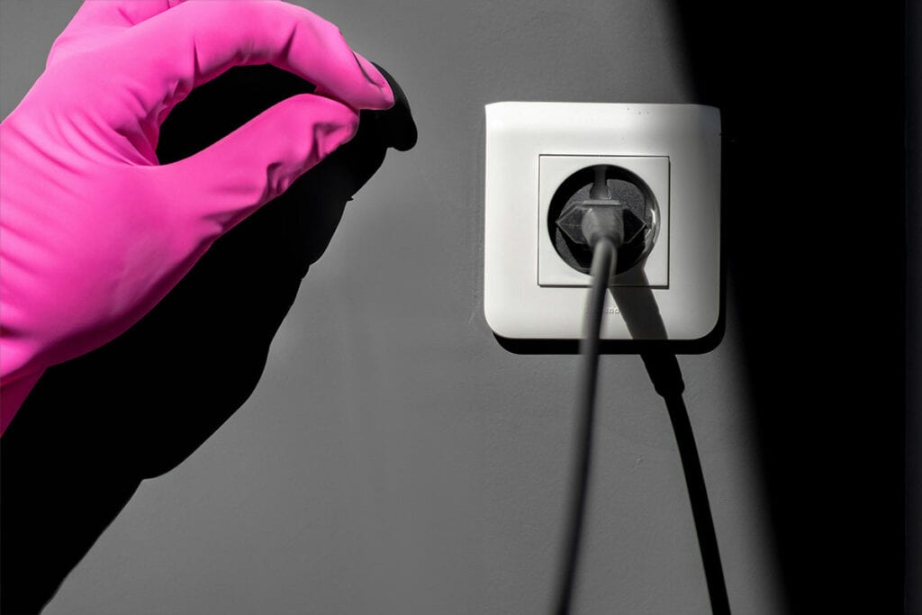 Eine Hand mit punkem Gummihandschuh nähert sich einem schwarzen Stecker in einer weißen Wandsteckdose